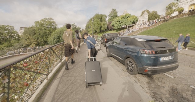 People walking along a path with luggage near Sacré-Cœur, Paris. Cars parked nearby, and love locks visible on the fence.