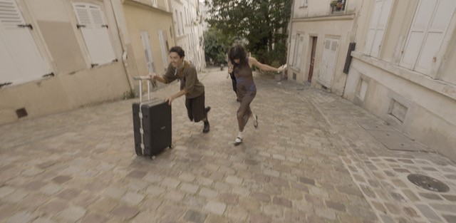 Two people rush down a cobblestone street with luggage in a narrow alley, surrounded by shuttered windows and aged buildings.