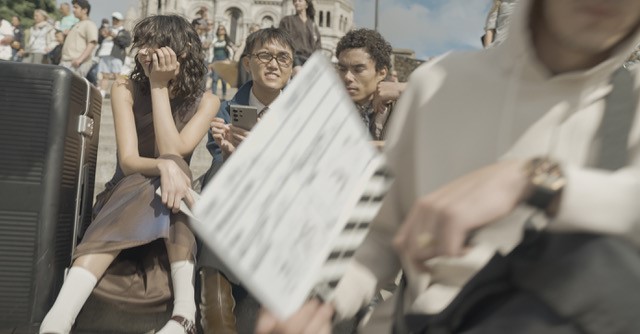 A group of people sit on steps, with one holding a clapperboard. In the background is the Sacré-Cœur Basilica, Paris.