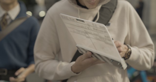 A person holds a movie clapperboard on a city street. Another person stands nearby. The scene appears busy and casual.