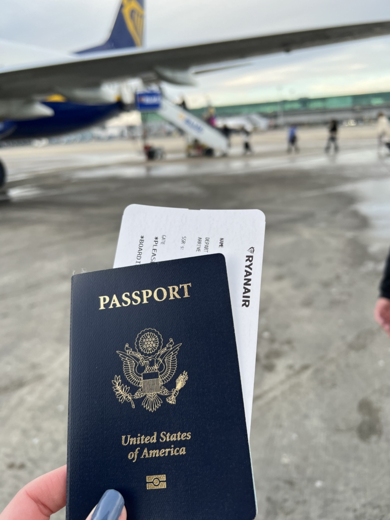 A person holds a U.S. passport and Ryanair boarding pass near an airplane at an airport. Other travelers and terminal visible.