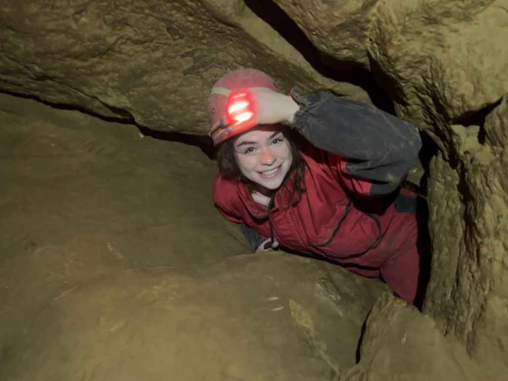 A person in a red jumpsuit with a helmet crawls through a narrow cave tunnel, smiling and adjusting the headlamp.