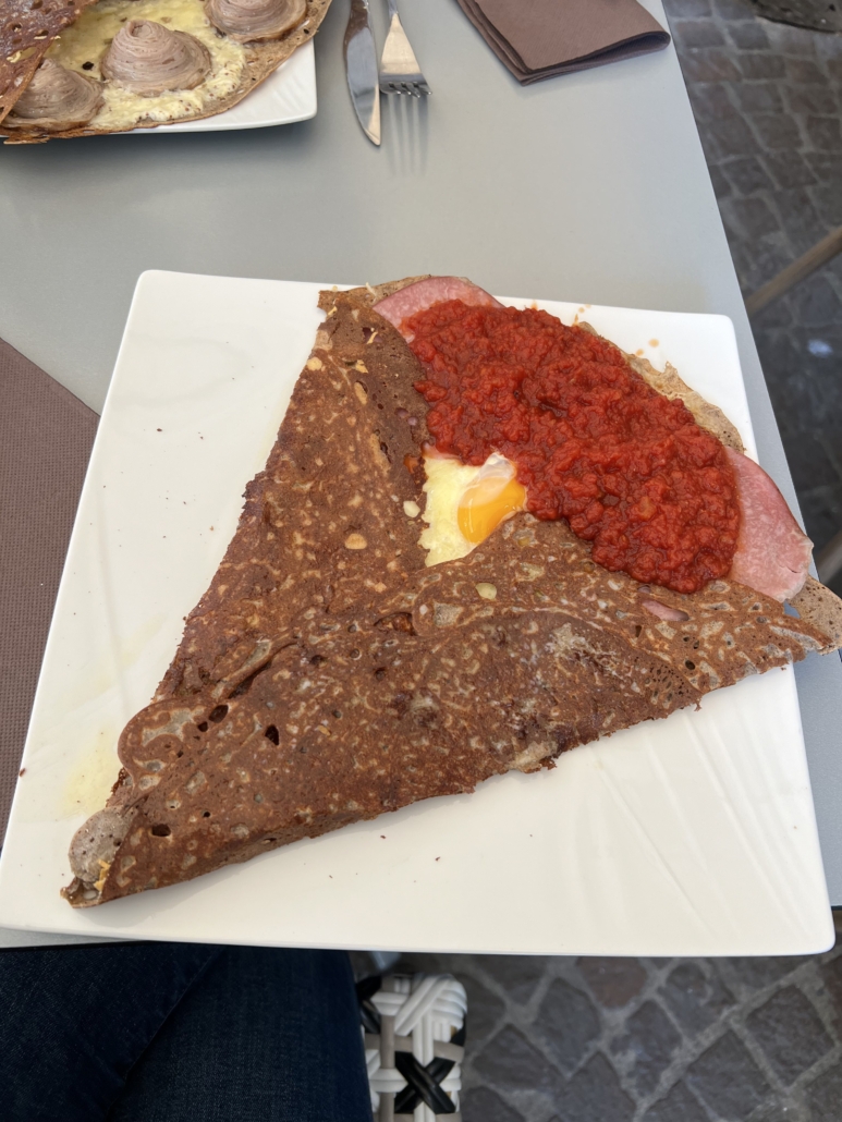 Buckwheat crepe with ham, egg, and tomato sauce on a square plate. Fork and knife nearby, outdoor setting with cobblestone ground.