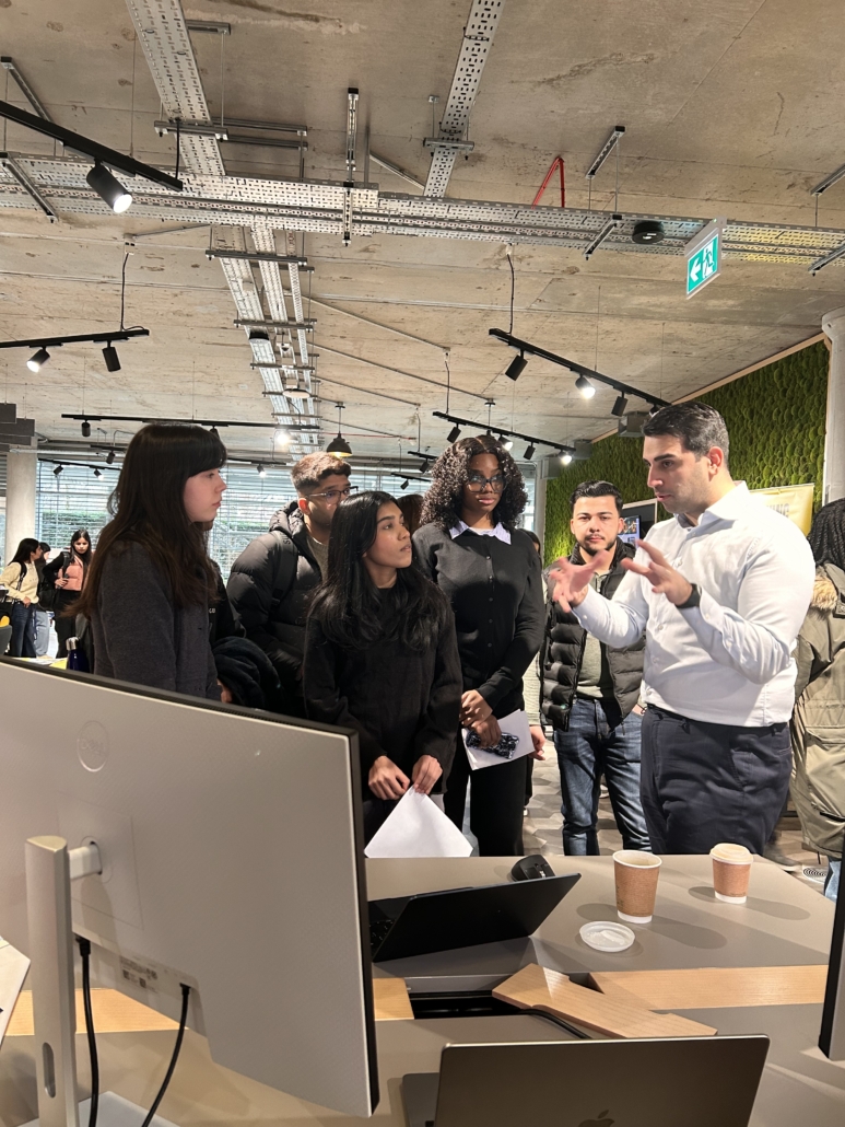 A person speaks to a group in a modern office setting, with computers and coffee cups on the table in front.