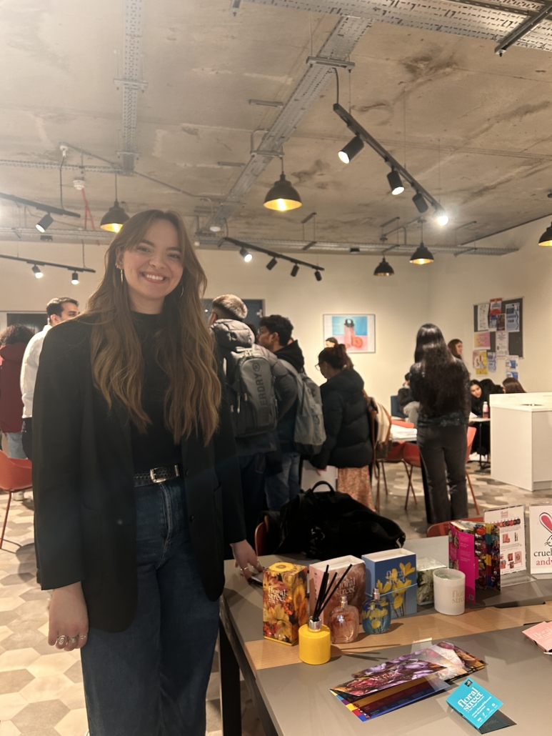 A person smiling in a busy, modern indoor space featuring tables with various colorful products and pamphlets on display.