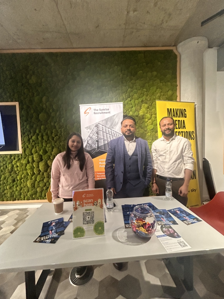 Three people at a recruitment event stand behind a table with brochures and candy, in front of green wall and promotional banners.