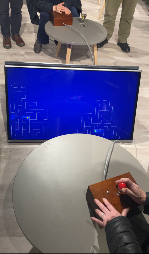 Person using a wooden joystick to play a maze game on a screen. Another person watches, with a glass on a table nearby.