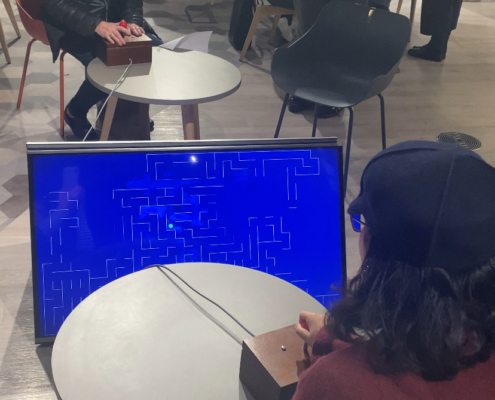 People are gathered indoors, engaging with a retro video game displayed on a screen. Tables and chairs are scattered throughout the room.