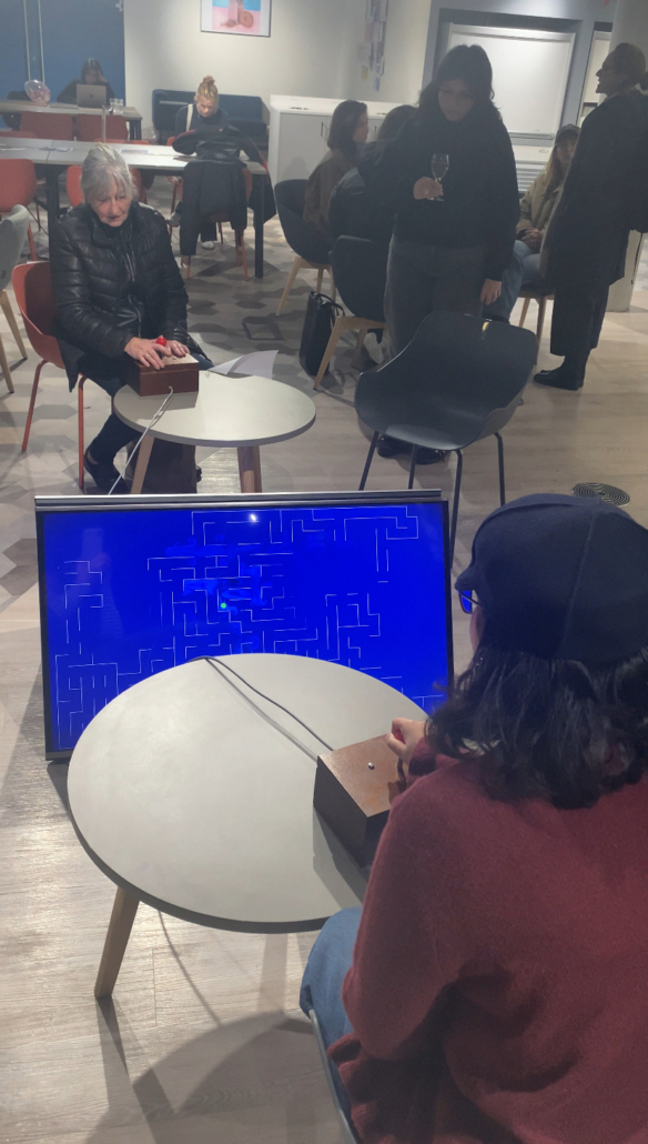 People are gathered indoors, engaging with a retro video game displayed on a screen. Tables and chairs are scattered throughout the room.