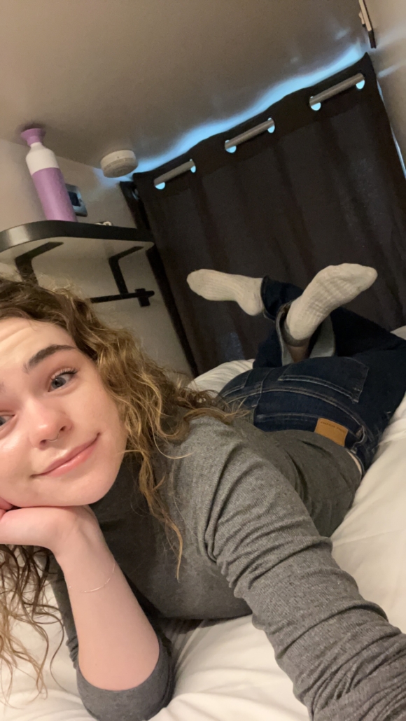 Person with curly hair lies on bed, smiling, wearing jeans and sweater. Purple bottle and smoke detector visible on shelf in cozy room.
