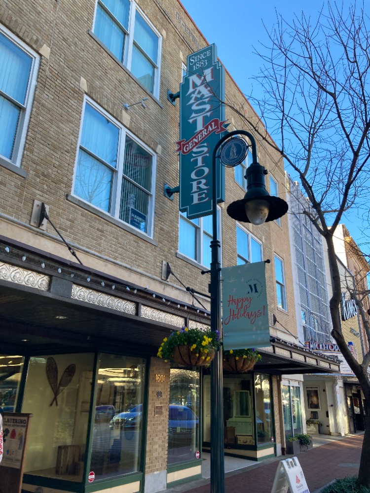 Street view of Mast General Store, historical building with signage and holiday decorations. A tree and a brick sidewalk are visible nearby.