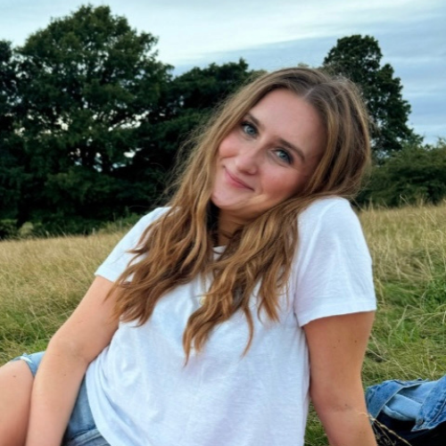 A person with long hair sits on grass wearing a white shirt with trees in the background, enjoying a natural setting on a sunny day.