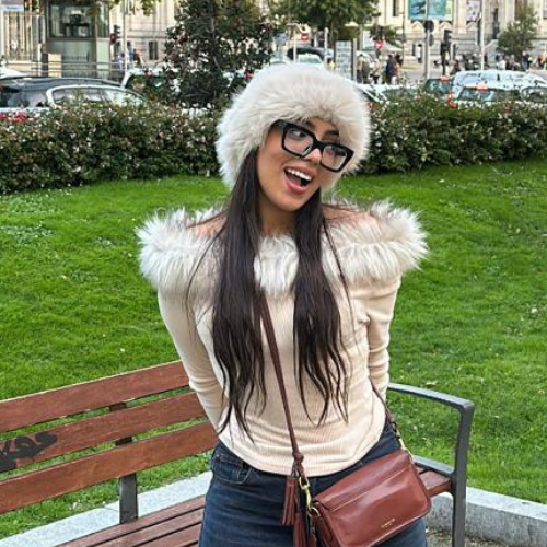 A person wearing glasses and a furry hat poses joyfully on a park bench with greenery and urban background, possibly in a city square.