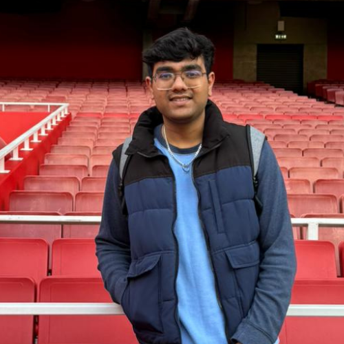 A person stands in front of red stadium seats at Emirates Stadium, wearing a blue and black jacket, smiling at the camera.