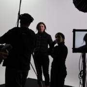 Three people in a studio setting with camera equipment and lighting; one is visible on a monitor screen.