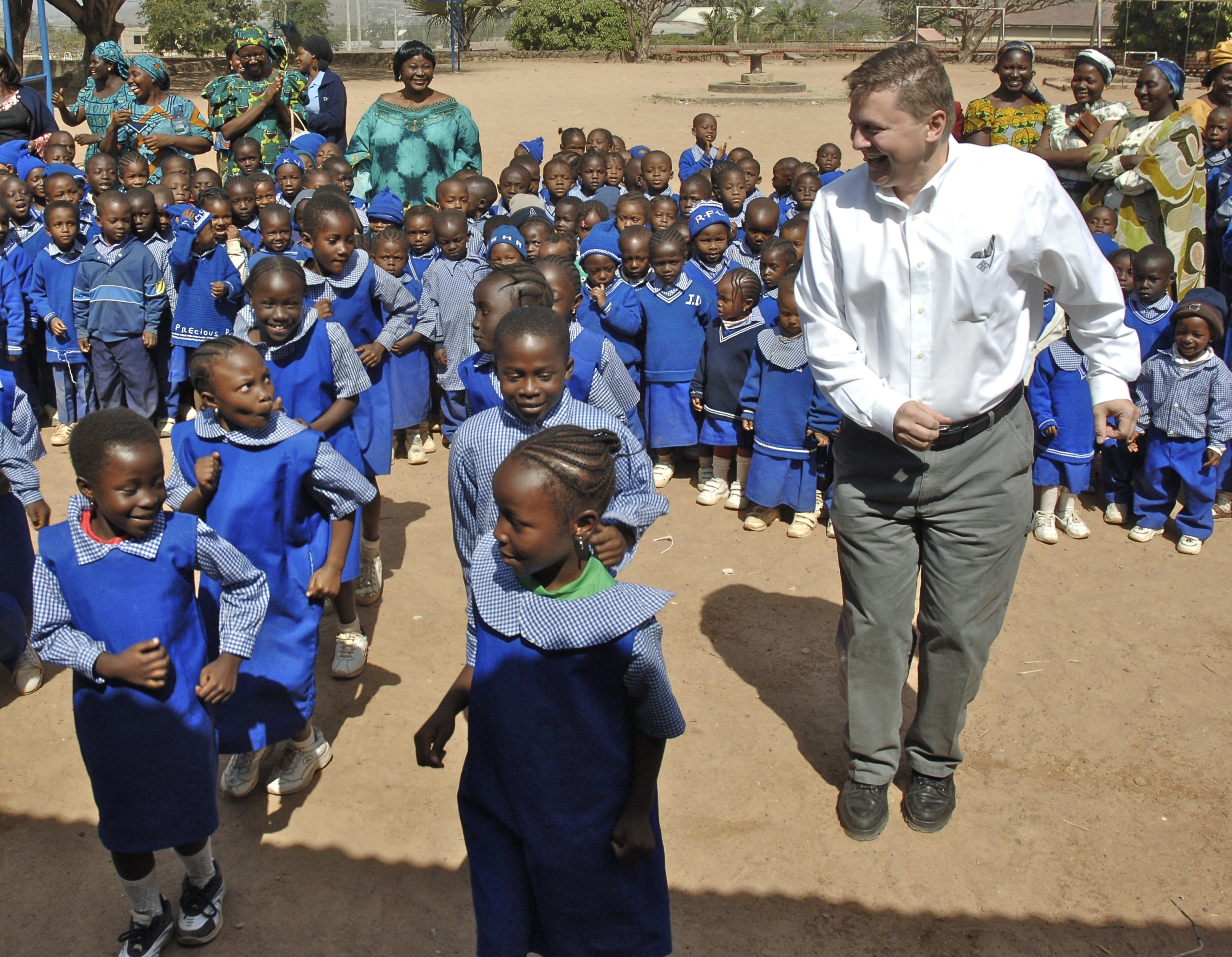 A group of children in blue uniforms dancing outside with a person in a white shirt, surrounded by smiling adults.