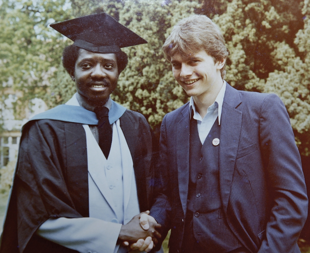 Two people shaking hands, one in a graduation gown, outdoors with trees in the background. Both appear cheerful, commemorating an academic achievement.