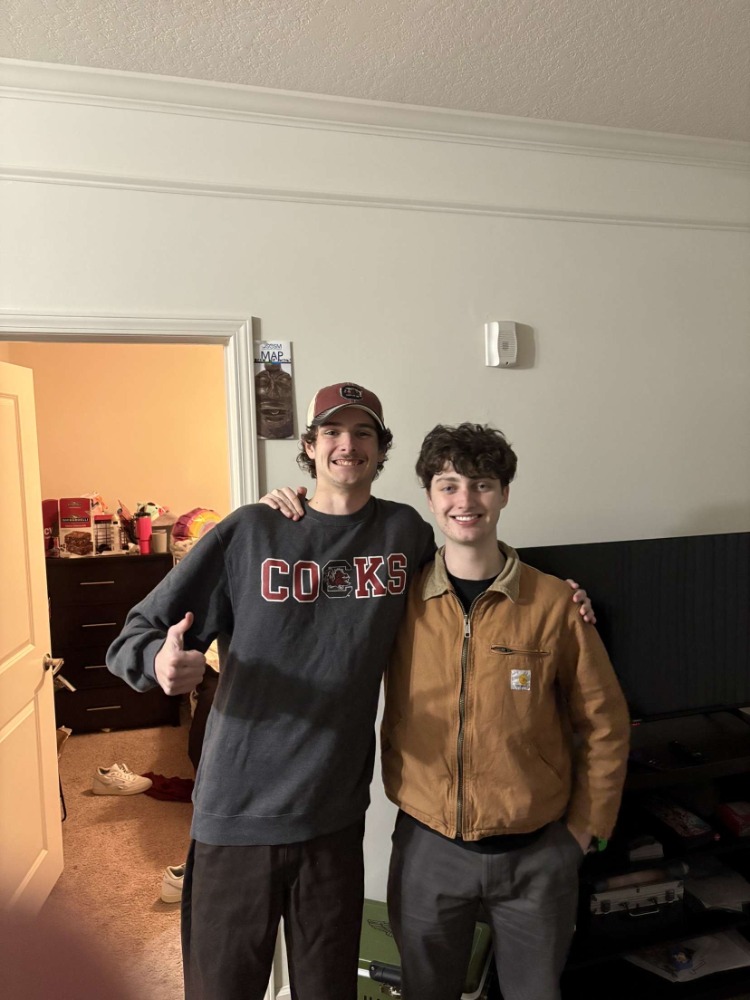 Two people smiling and posing in a room with an open doorway revealing a cluttered interior. A poster is partially visible on the wall.