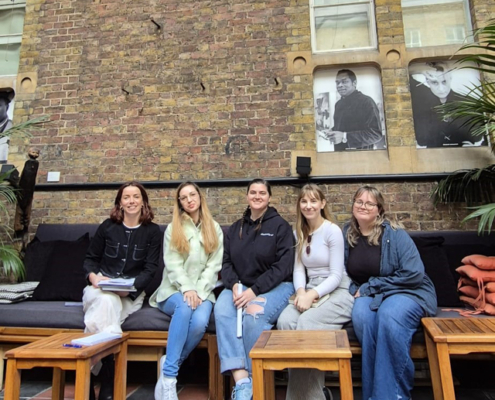 Five people sitting on a bench against a brick wall with arched windows and black-and-white portraits, surrounded by tables and plants.