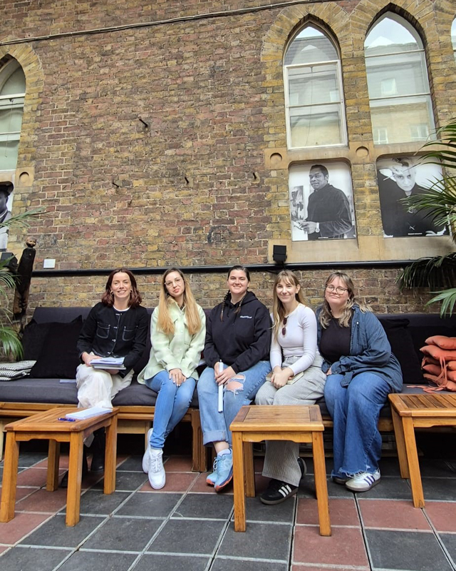 Five people sitting on a bench against a brick wall with arched windows and black-and-white portraits, surrounded by tables and plants.