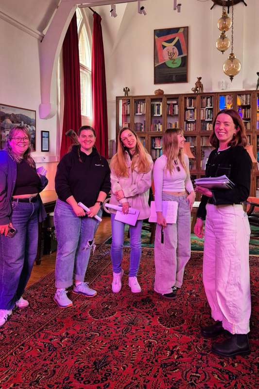 Five people stand in a room with bookshelves and art. They appear to be in a historical building, engaged in discussion.