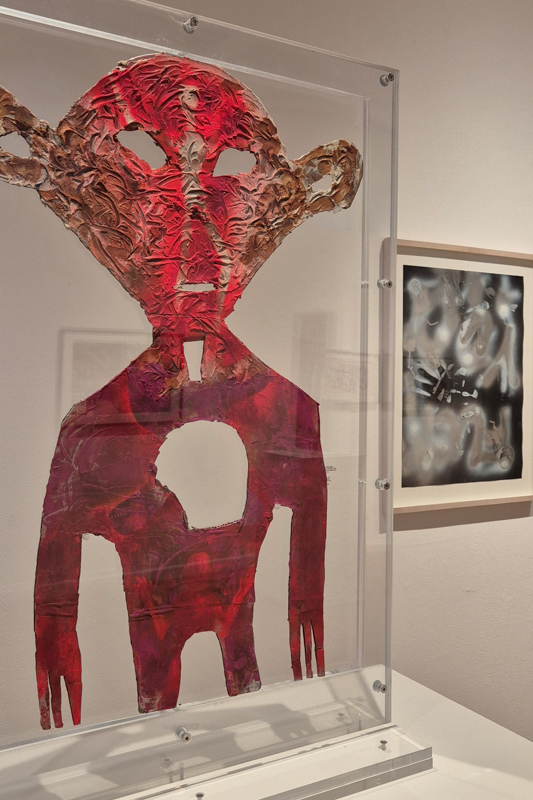 Abstract red figure artwork in a glass case, displayed on a white stand in a gallery. A blurred framed piece is visible behind it.
