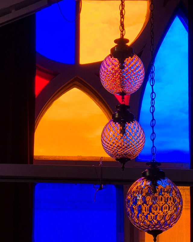 Colorful stained glass windows illuminate decorative hanging lamps, casting warm, vibrant hues, creating a cozy and enchanting atmosphere in an indoor space.