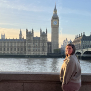 A person stands by the River Thames with the iconic Big Ben and Houses of Parliament in the background, visible under clear skies.