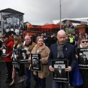 People holding "Justice" signs with photos, walking in front of Bogside mural, demanding accountability. Cloudy sky, buildings in the background.