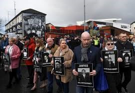 People holding "Justice" signs with photos, walking in front of Bogside mural, demanding accountability. Cloudy sky, buildings in the background.