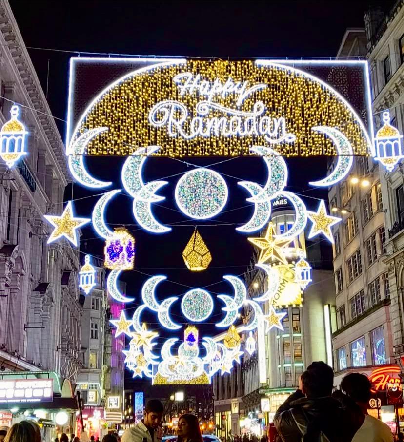 Street with illuminated "Happy Ramadan" decorations, featuring crescents and stars. People walk beneath festive lights in a bustling urban setting.