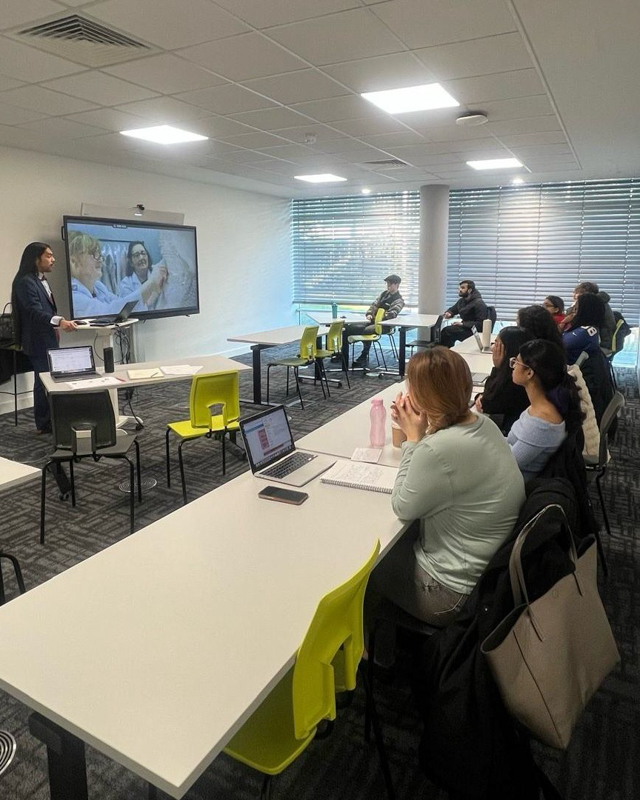 A person presents to classroom attendees, discussing a topic on-screen. Attendees are seated at desks with laptops and notebooks in a modern room.