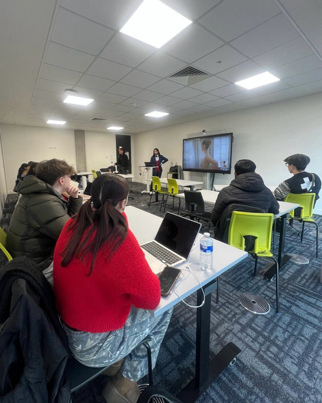 People in a classroom setting with a presentation displayed on a large screen. Several attendees are using laptops at desks.