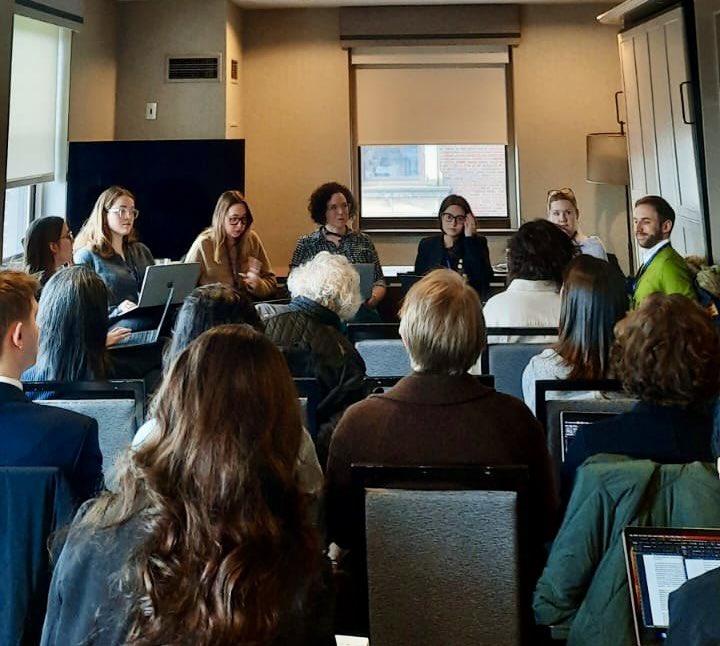 A group of people sits in a conference room during a presentation or meeting, with laptops visible and window in the background.