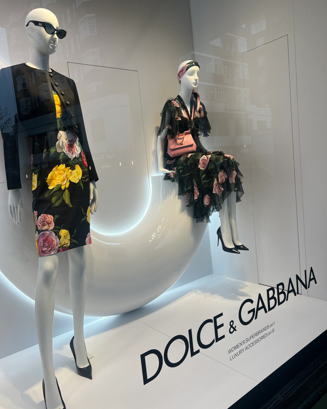Two mannequins in floral dresses are displayed in a Dolce & Gabbana storefront, featuring stylish accessories and a modern background design.