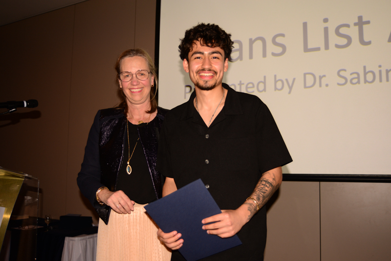 Two people smiling at an awards ceremony, holding a certificate. A presentation screen displays text in the background.