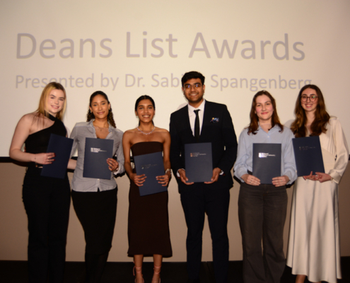 Six persons smile holding certificates in front of a projected screen at the Deans List Awards ceremony. Formal attire is worn.