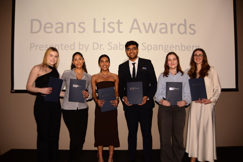 Six persons smile holding certificates in front of a projected screen at the Deans List Awards ceremony. Formal attire is worn.
