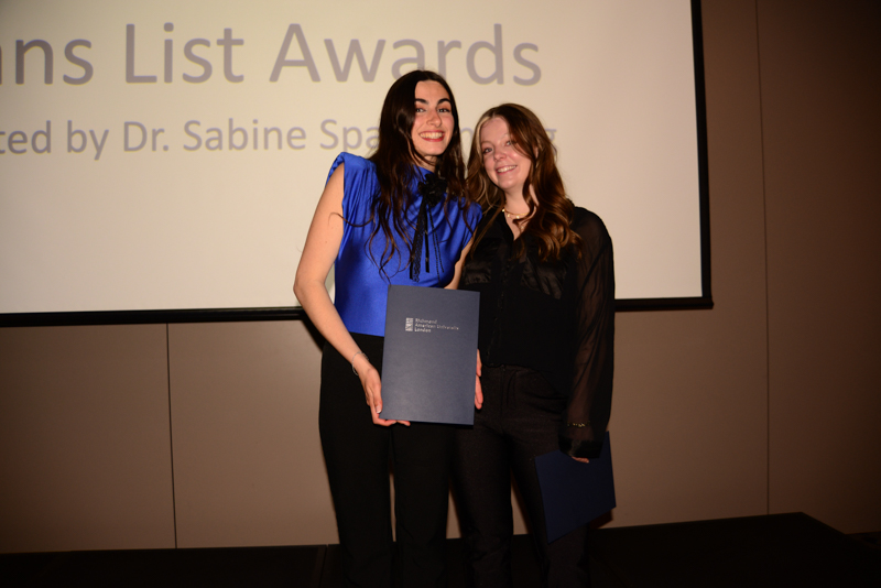 Two people pose with certificates at an awards ceremony. A projection in the background partially reads "List Awards," indicating an academic event.
