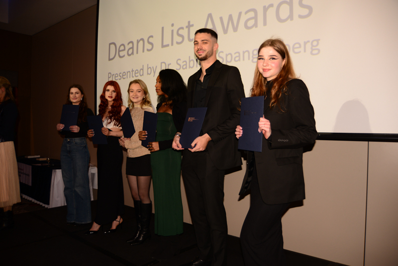 Six people standing on stage holding certificates, posing for a photo at an awards event with a presentation screen in the background.