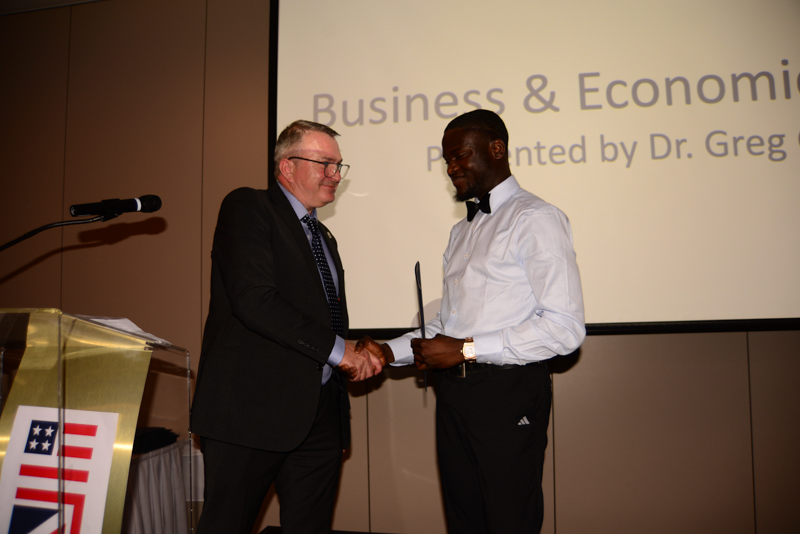 Two people are shaking hands at a business and economics presentation. A podium with a flag design is visible.