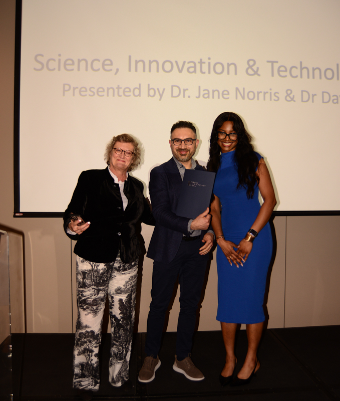 Three people standing on stage smiling, holding a certificate. Behind them, a presentation screen reads "Science, Innovation & Technology" with presenters' names.