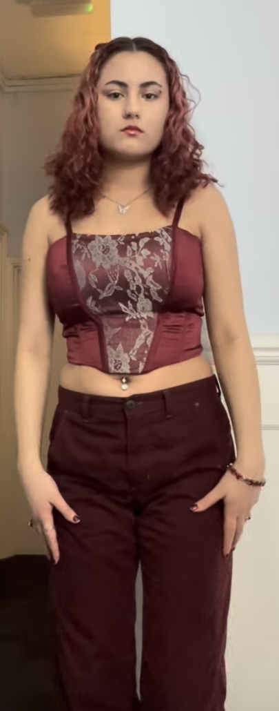 Person with curly hair in a maroon lace top and pants, standing indoors with neutral expression and light blue background.
