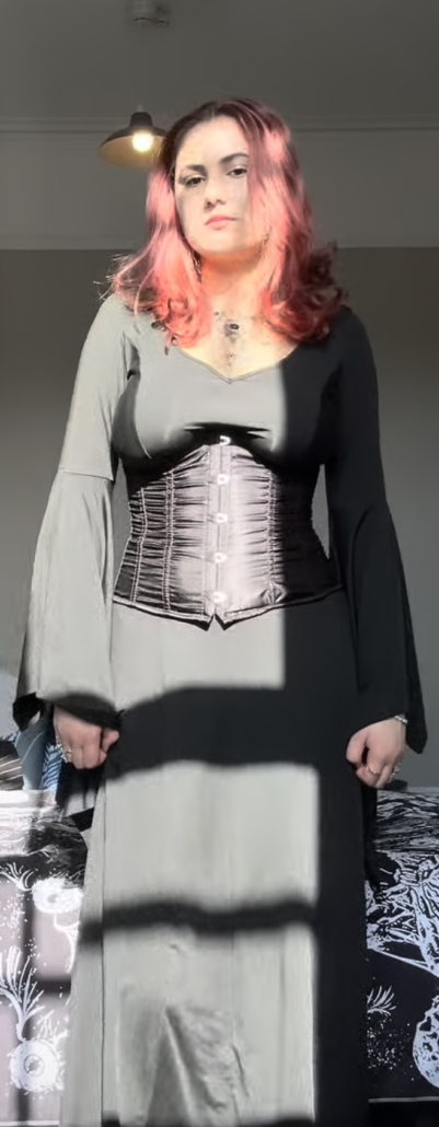A person with dyed pink hair stands indoors, wearing a long dress and corset, sunlight casting shadows on them.
