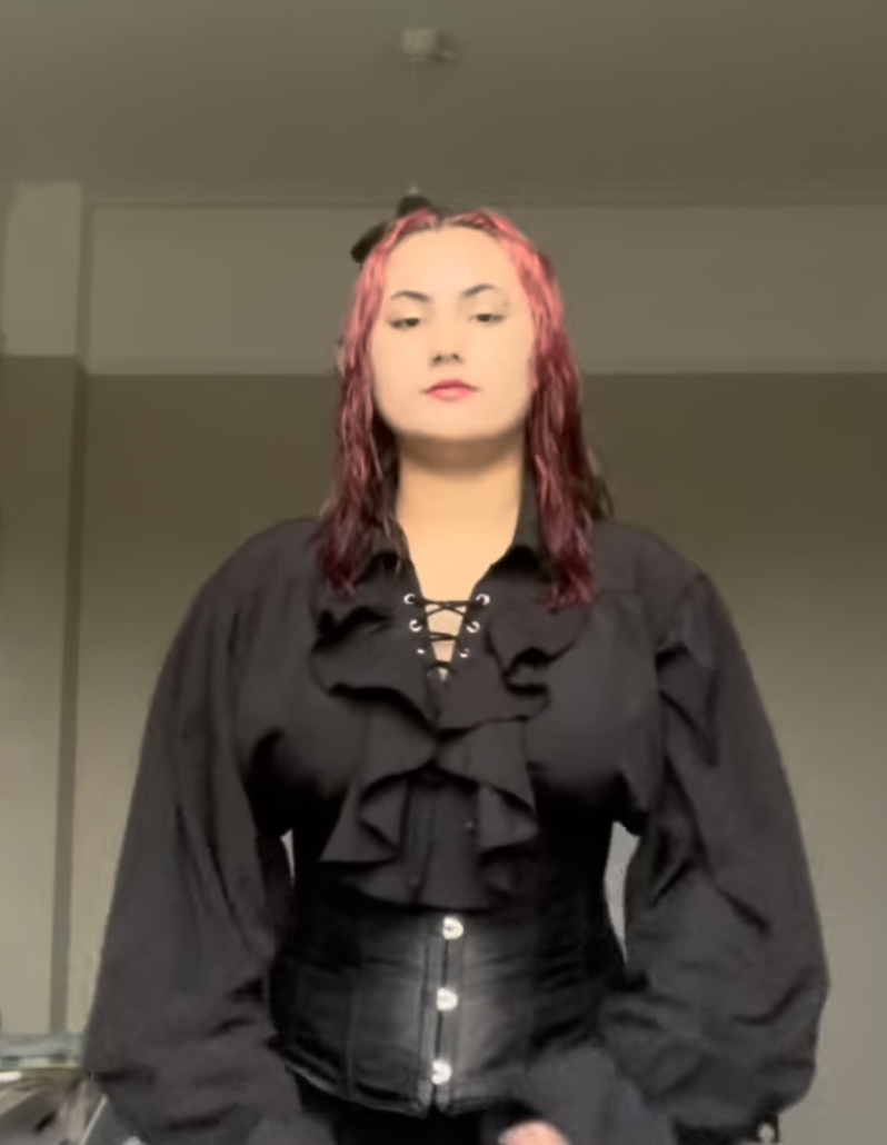 A person with dyed hair wearing a black lace-up blouse and corset stands indoors, against a plain background, looking directly at the camera.