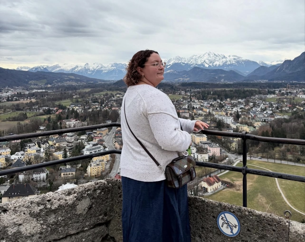A person stands on a terrace at Hohensalzburg Fortress, overlooking Salzburg's picturesque landscape with distant snow-capped mountains under a cloudy sky.