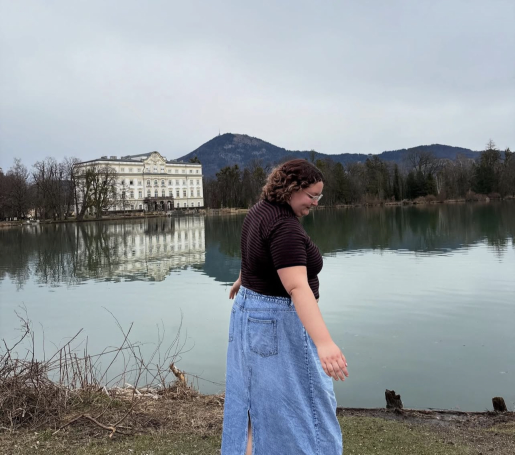 A person walks by a lake with Schloss Leopoldskron reflecting in the water, surrounded by trees and hills in the background.