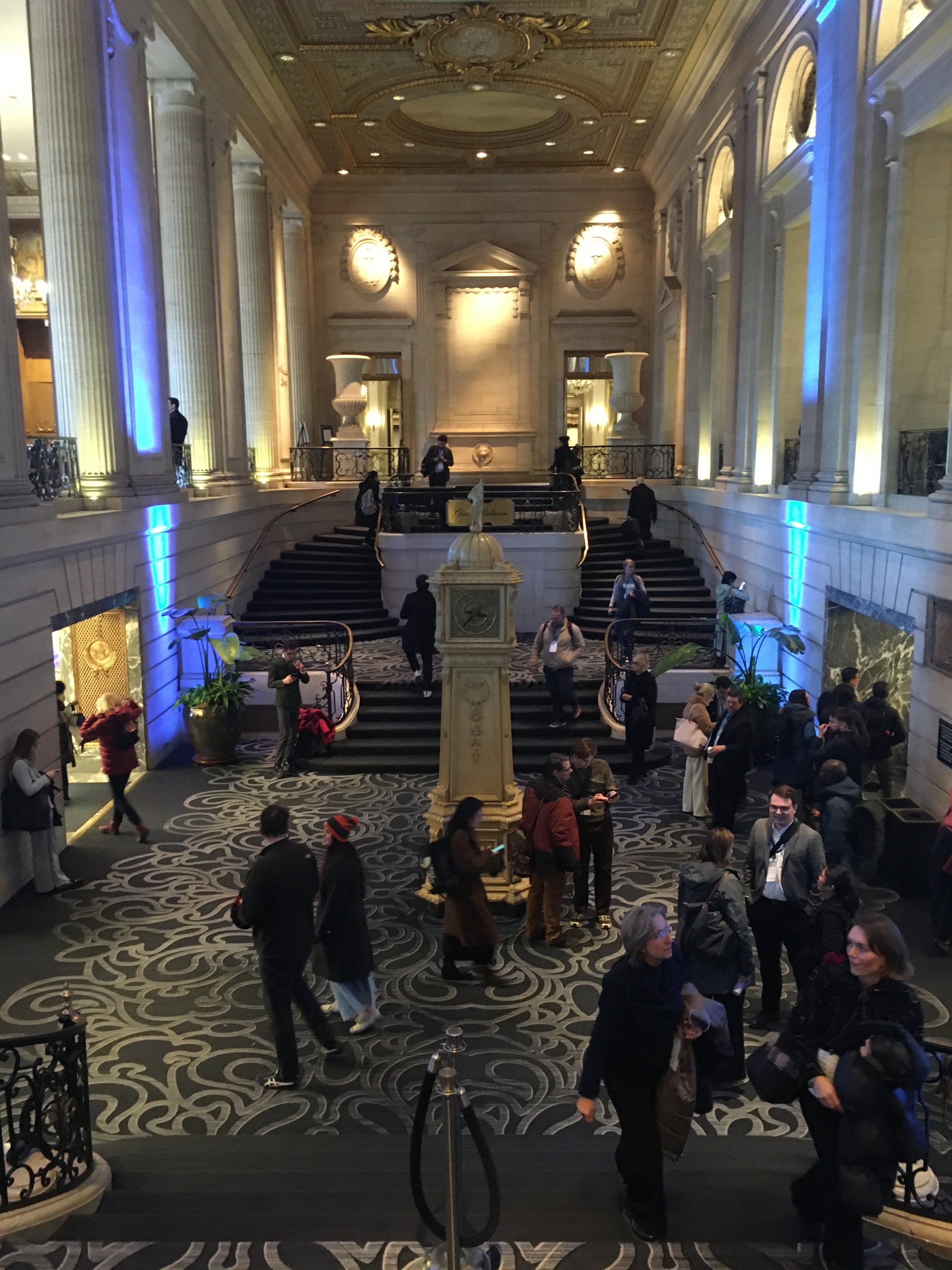 An elegant hall with ornate ceiling and columns, bustling with people on carpeted stairs, illuminated with blue lighting, suggests a grand event space.