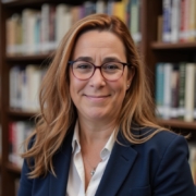 A person with glasses smiles in front of a bookshelf, wearing a blue blazer and white shirt, in a library setting.
