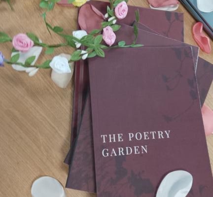 Stack of maroon "The Poetry Garden" books on a table with decorative flower petals scattered around, creating an artistic and inviting ambiance.